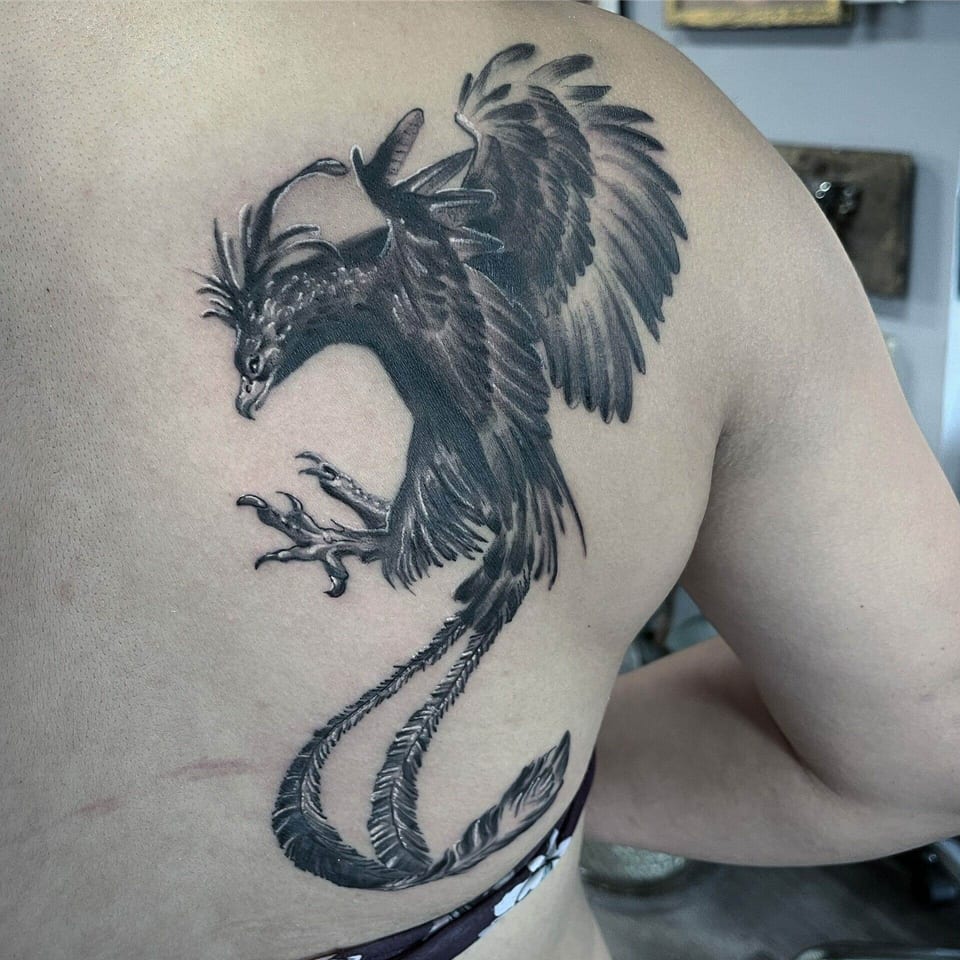 A black and grey winged dragon with a curling feathered tail covers a shoulder blade.