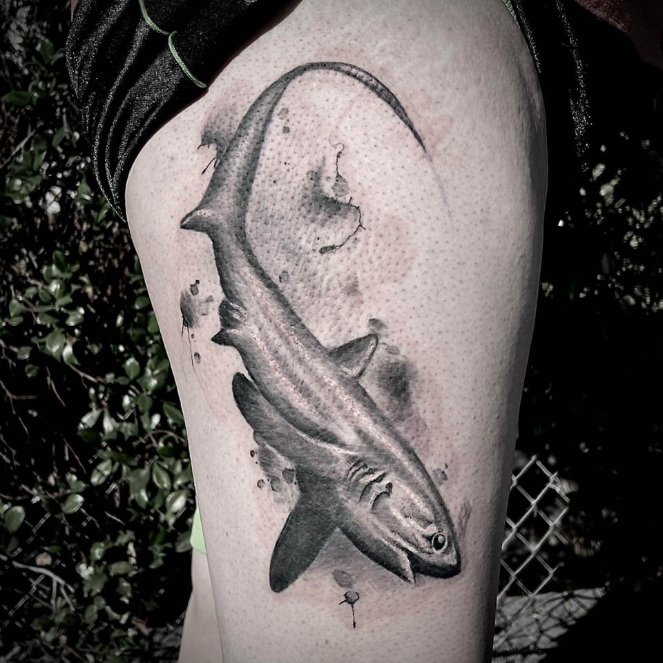 A black and grey realism shark tattoo curves down the outer thigh.