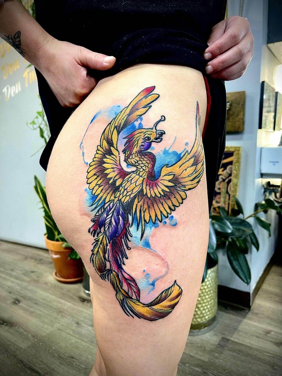 A golden illustrative phoenix spreads its wings over blue watercolor splashes on a thigh.