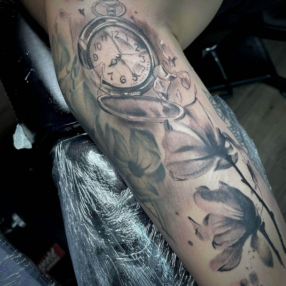 A black and grey pocket watch with broken glass and birds sits on an inner upper arm.