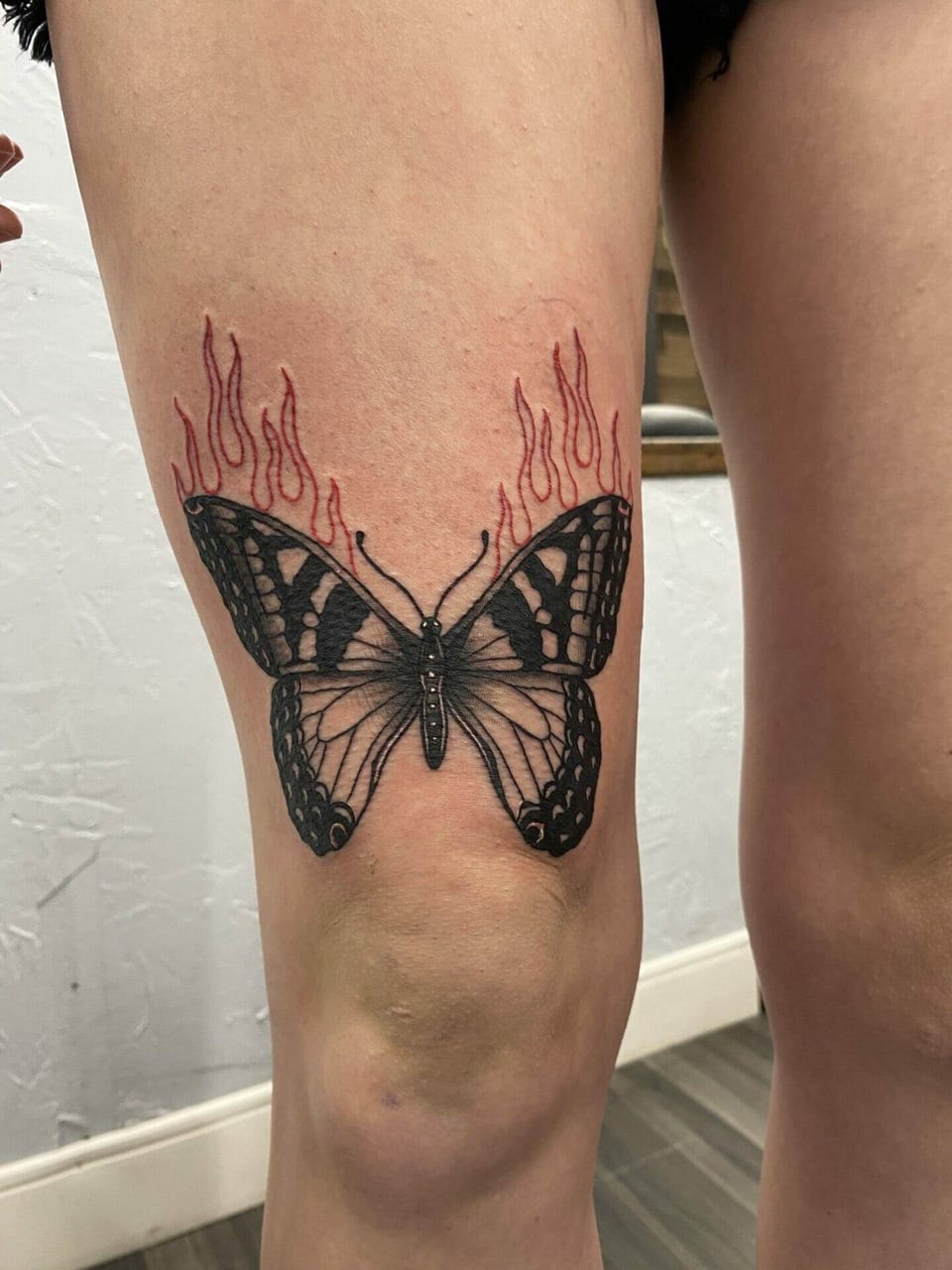 A blackwork butterfly with red flames sits above a knee.