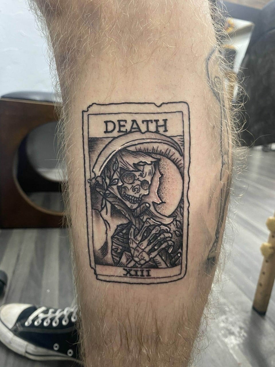 A black and grey Death tarot card with a skeleton and scythe sits on a lower leg.
