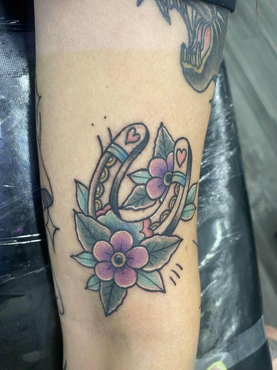 A traditional horseshoe with hearts and purple flowers sits on a forearm.