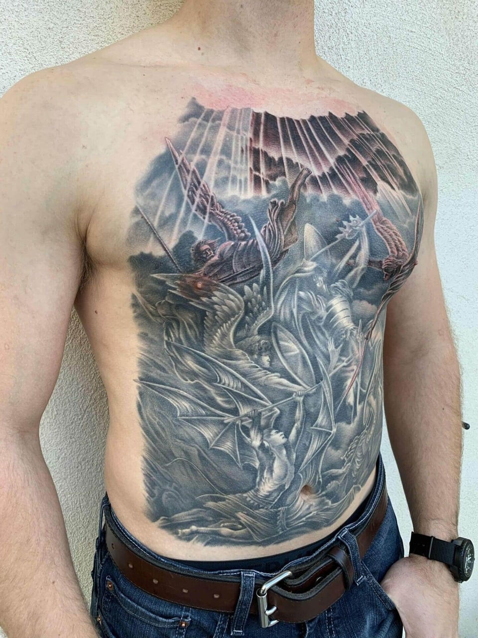Angled view of a black and grey battle of knights, angels and a dragon across the chest and stomach, with a wristwatch.