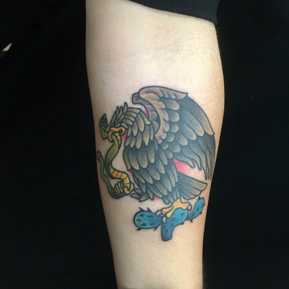 A traditional eagle grips a green snake above a blue cactus on the forearm.