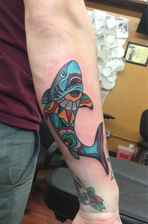 A traditional shark with blue fins and a multicolored stained-glass body curves along the forearm.