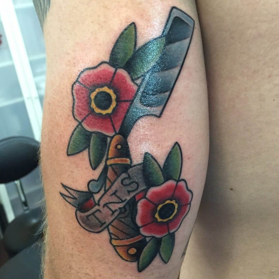 A traditional straight razor sits between red flowers and green leaves with a banner on the upper arm.