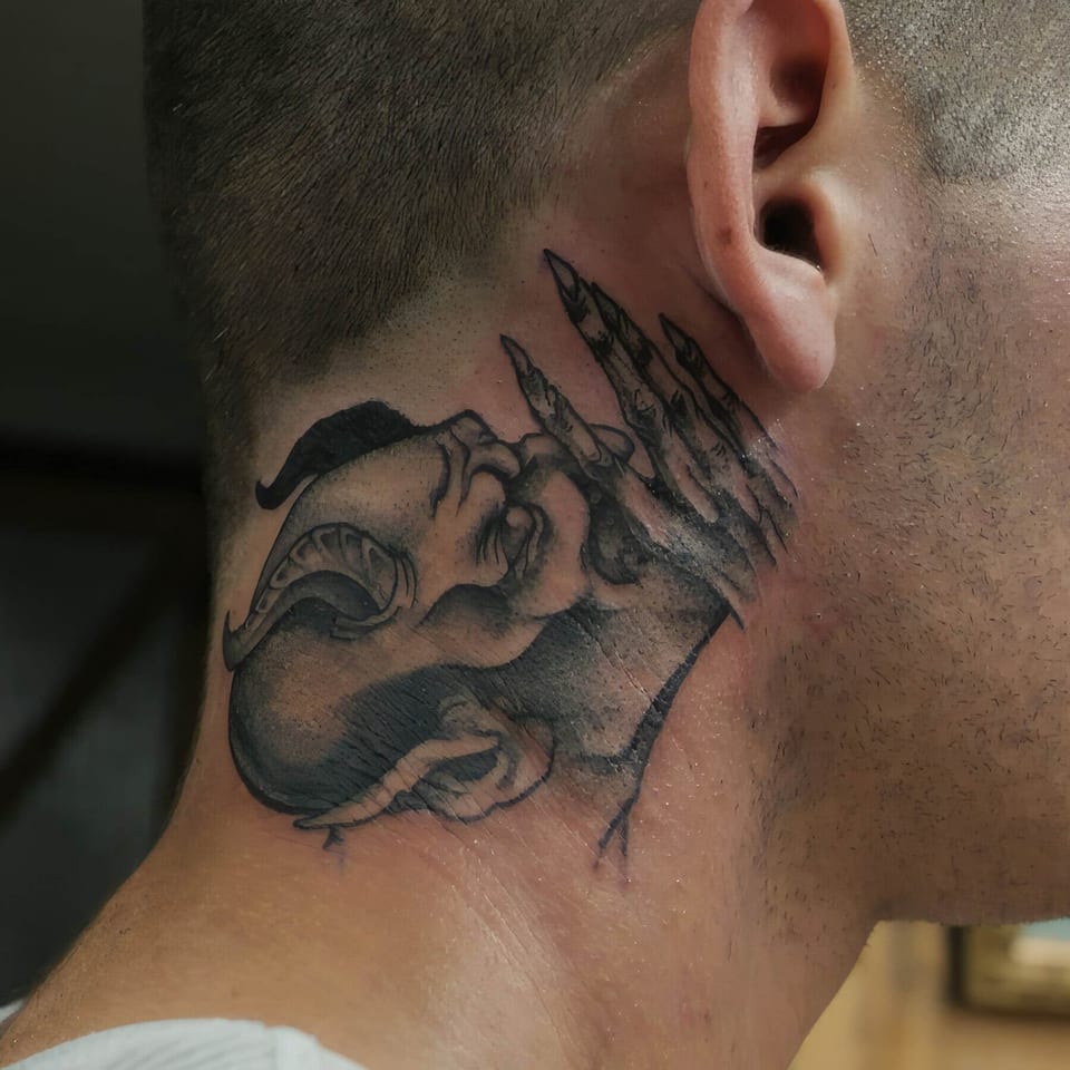 A black and grey horned devil with a raised clawed hand sits behind the ear on the neck.