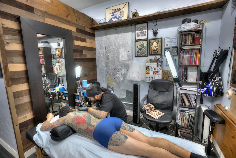 An artist tattoos a client's Japanese back piece beside mirrors, framed artwork and book-filled shelves.