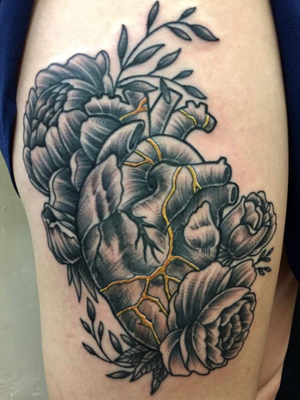 A black and grey anatomical heart with gold cracks sits among peonies and leaves on the upper arm.