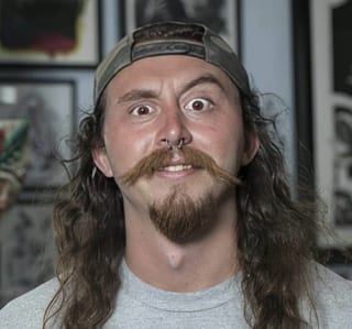 Hunter Knolmayer faces the camera wide-eyed with long hair, a mustache and a backward cap.
