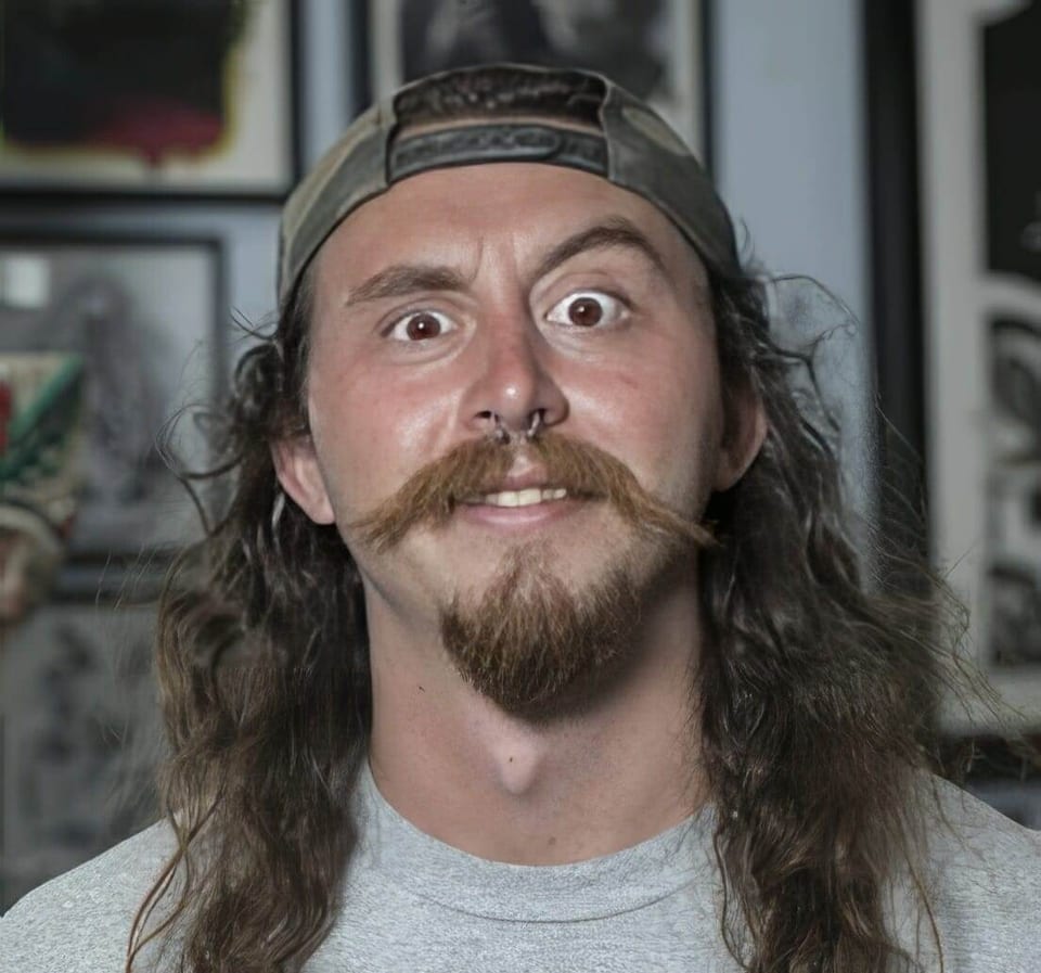 Hunter Knolmayer faces the camera wide-eyed with long hair, a mustache and a backward cap.