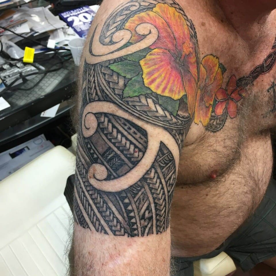 A color hibiscus and red plumeria sit above black Polynesian bands on a man's shoulder and chest beside shop computers.