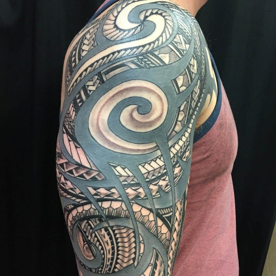 A black and grey Polynesian shoulder tattoo combines large spirals, angular bands and hexagonal patterns.