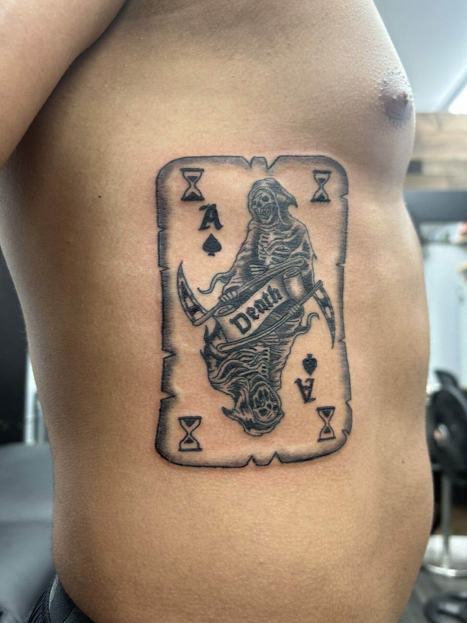 A black and grey playing card tattoo places a mirrored grim reaper and hourglasses on the ribs.