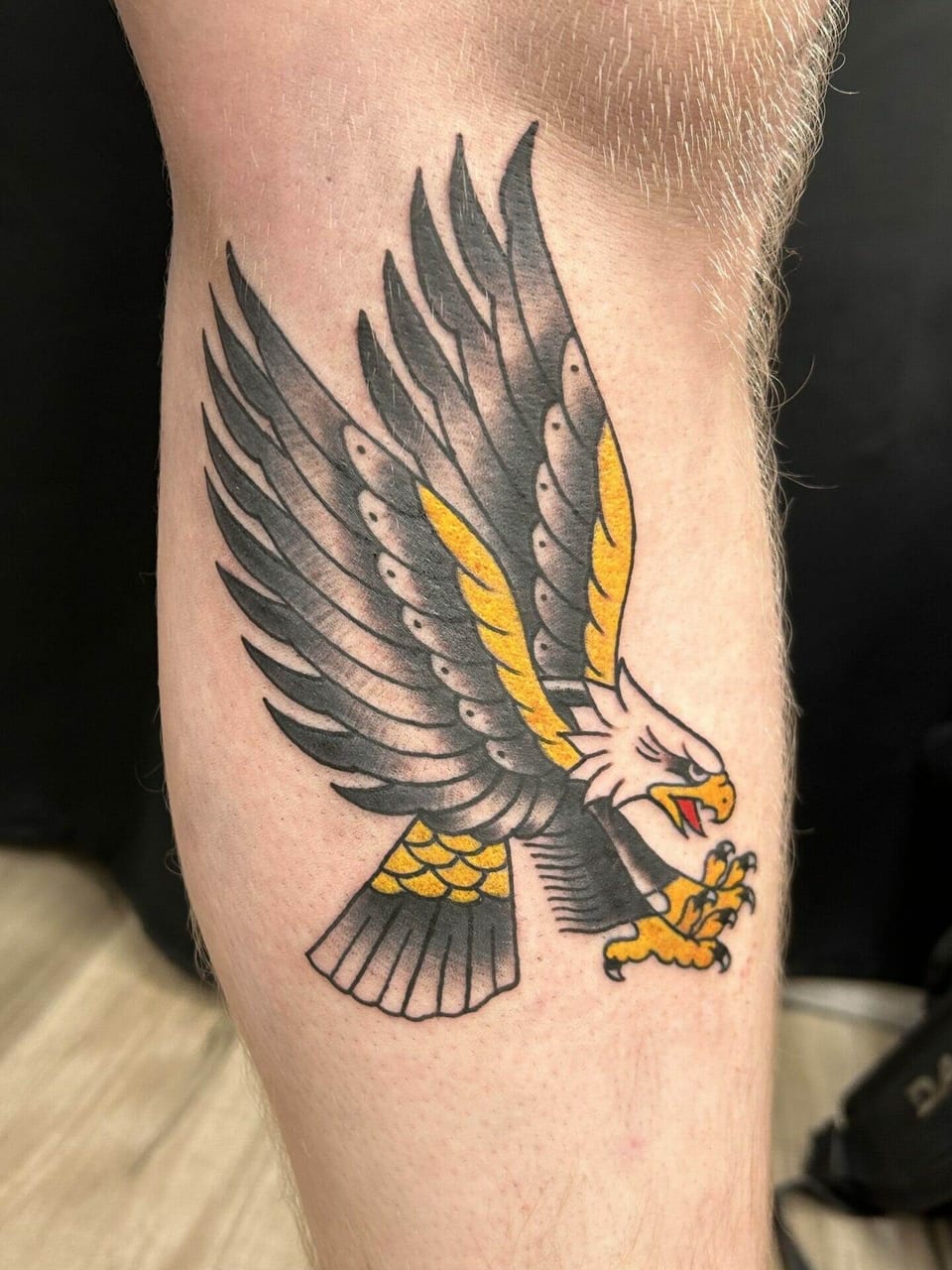 A traditional eagle tattoo spreads black and grey wings with yellow accents across a lower leg.