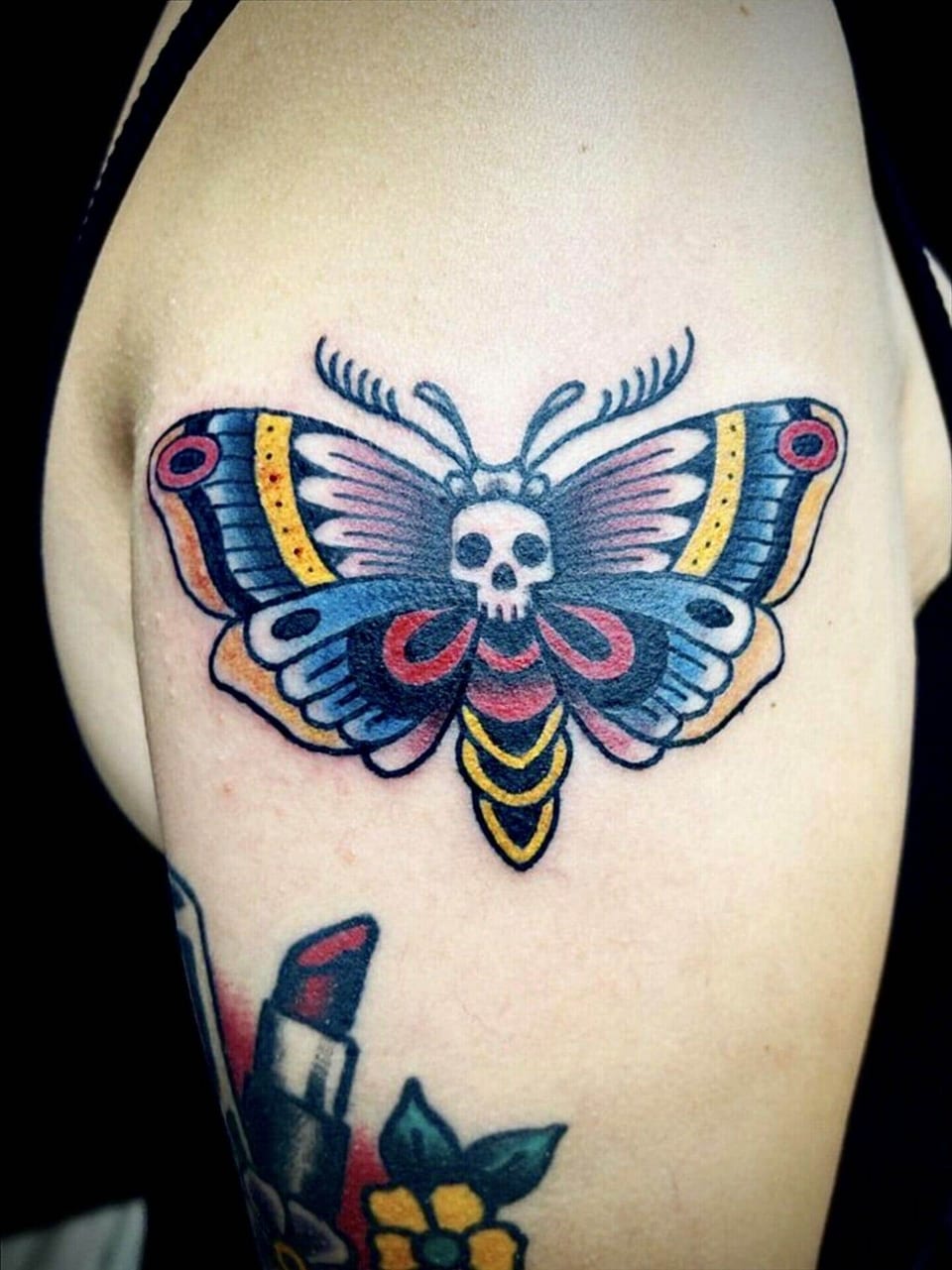 A traditional death's-head moth tattoo spreads blue, red and yellow wings across an upper arm.