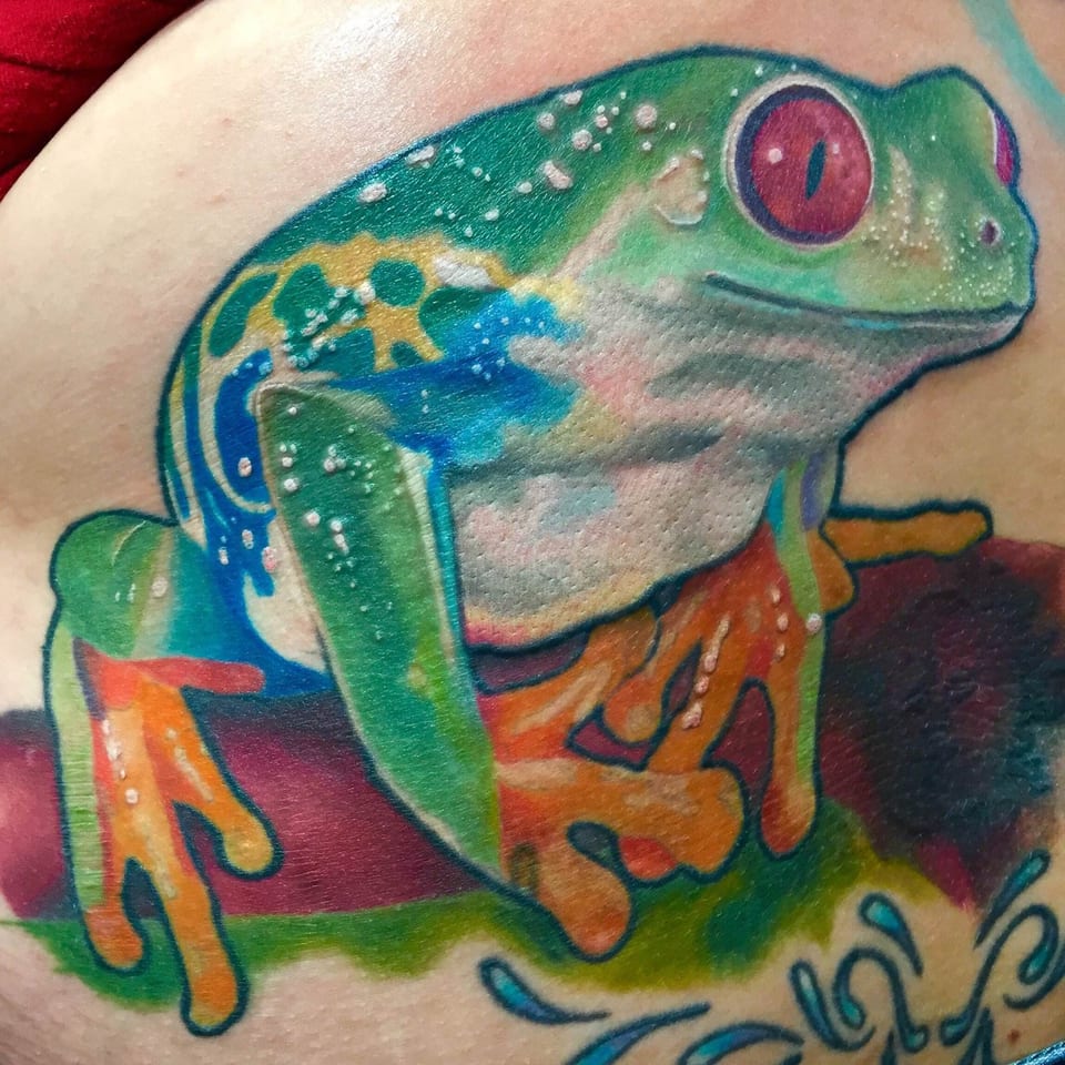 A color realism red-eyed tree frog tattoo rests on a branch across a thigh.