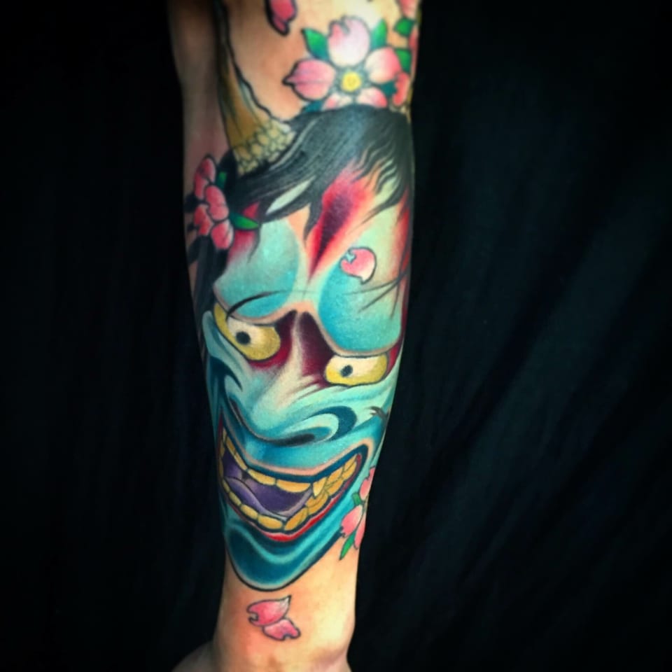 A Japanese blue hannya mask tattoo has yellow eyes and pink cherry blossoms on a forearm.