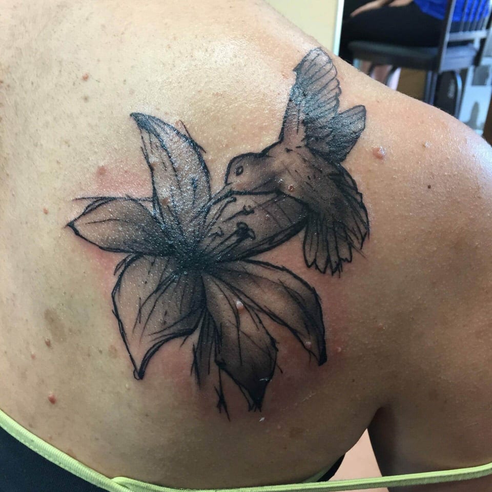 A black and grey illustrative hummingbird tattoo hovers beside a lily on a shoulder blade.