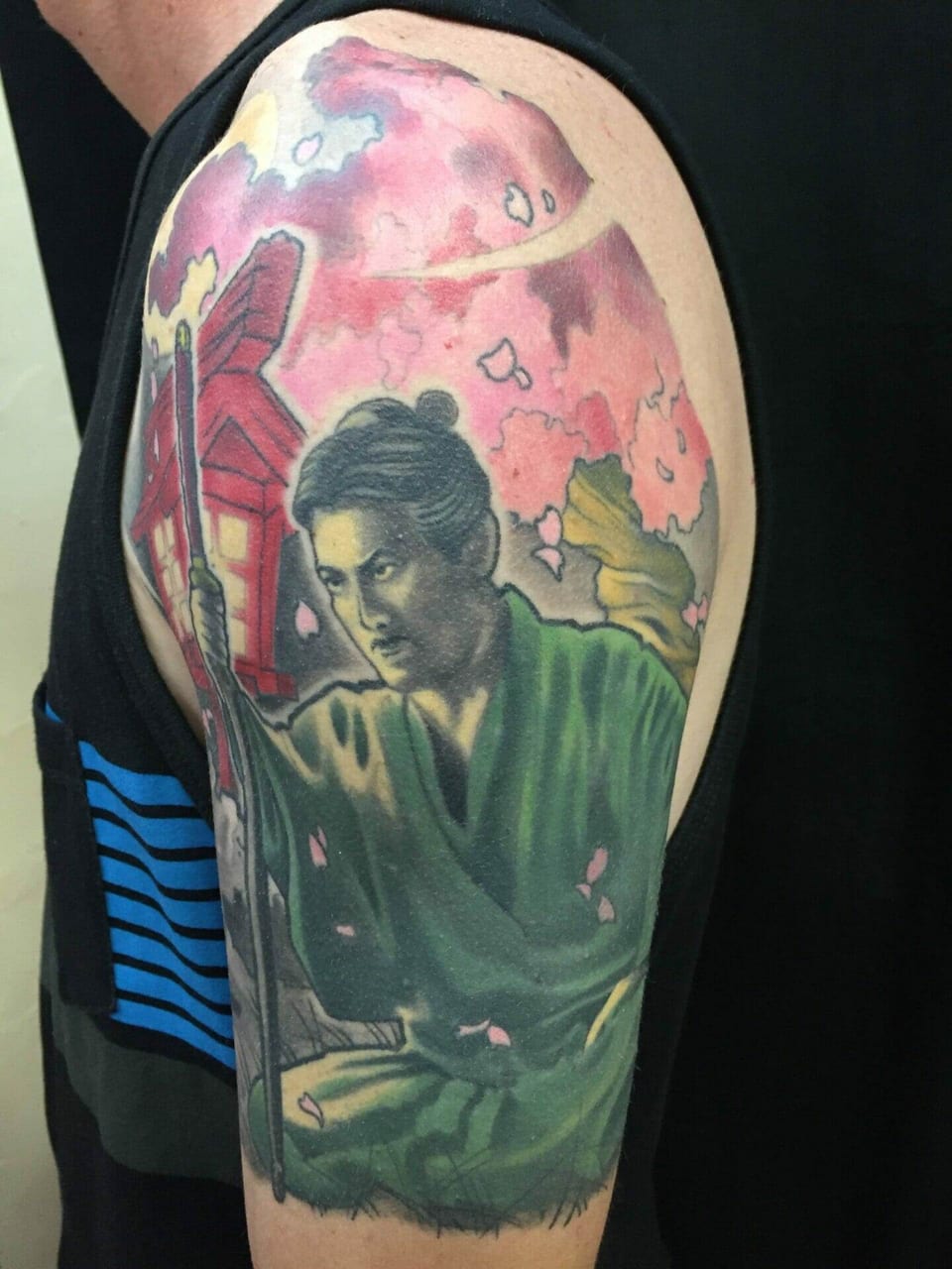 A Japanese samurai tattoo holds a sword beneath pink blossoms beside a red building on an upper arm.