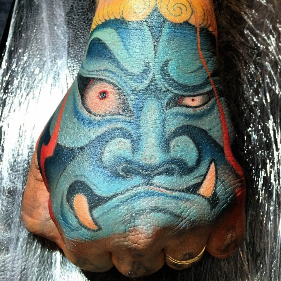 A Japanese blue oni mask tattoo bares orange fangs across the back of a hand.