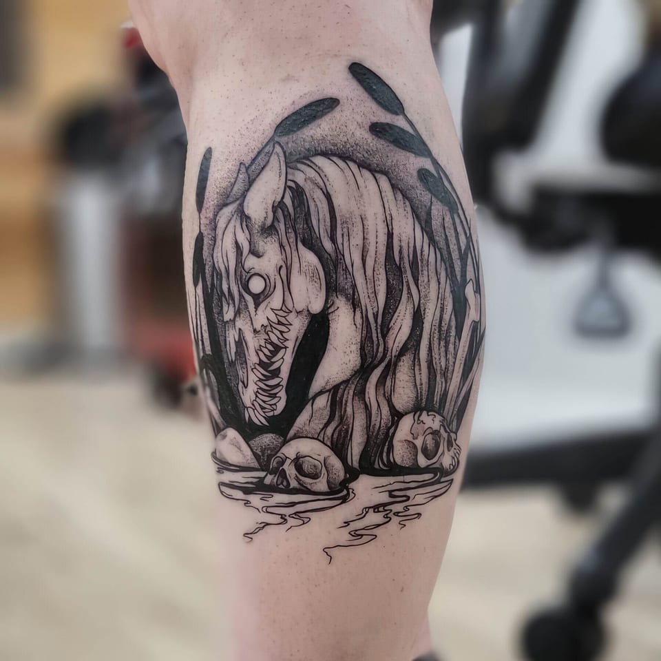 A black and grey kelpie rises from water with reeds and skulls around its flowing mane on a calf.