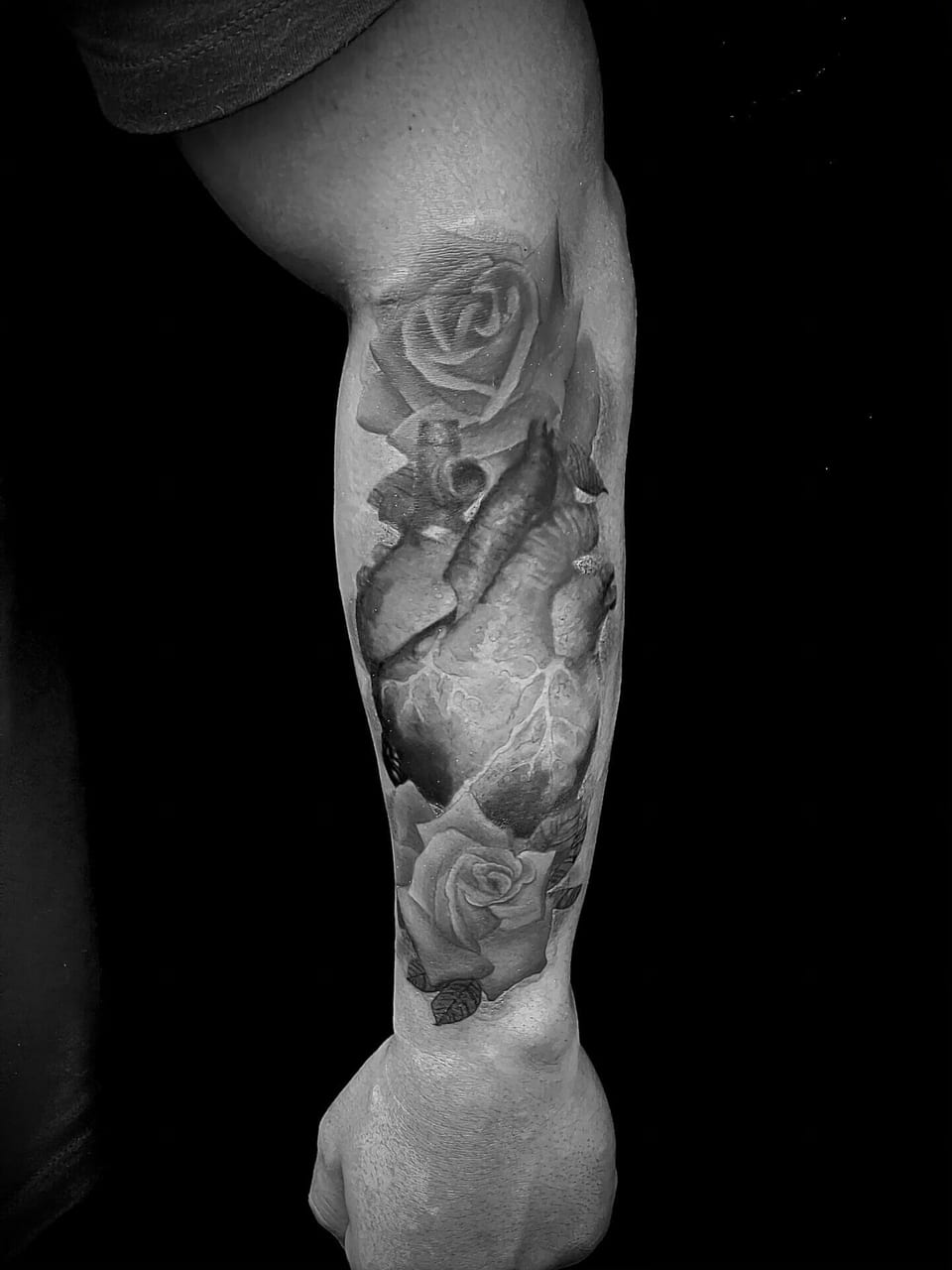 A realistic black and grey anatomical heart sits between roses along the forearm.