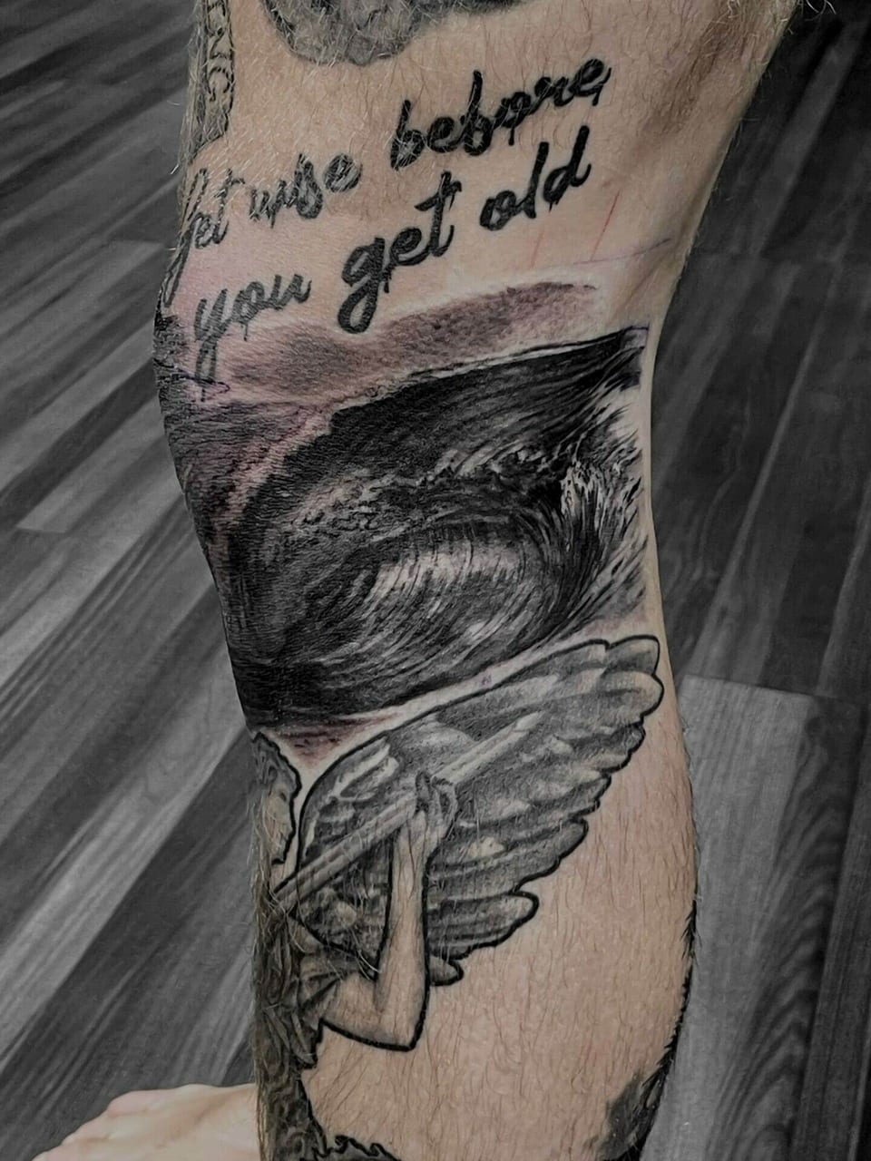 A black and grey ocean wave and winged figure cover a calf beneath a line of script.