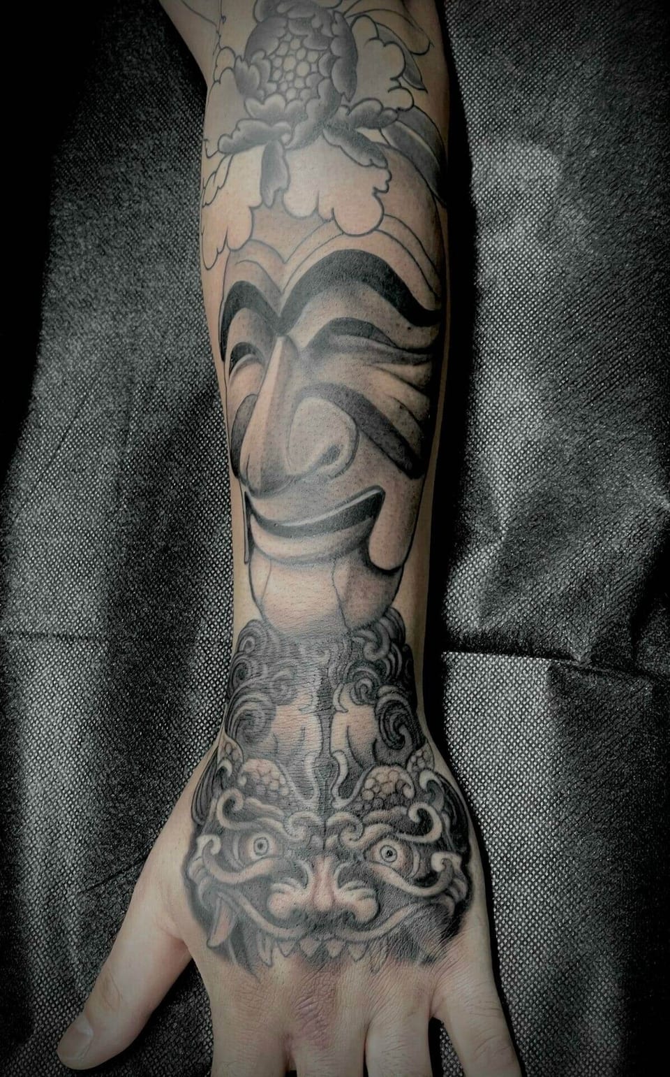 Black and grey Japanese masks, a guardian lion and a peony extend along a forearm onto the hand.