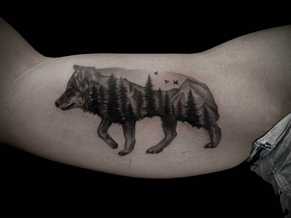 A black and grey wolf silhouette contains mountains, pine trees and flying birds on an upper arm.