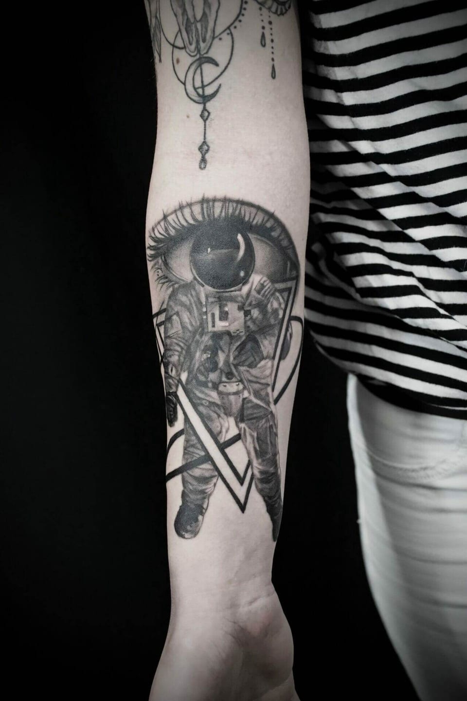 A black and grey astronaut floats beneath a large eye within triangular outlines on a forearm.