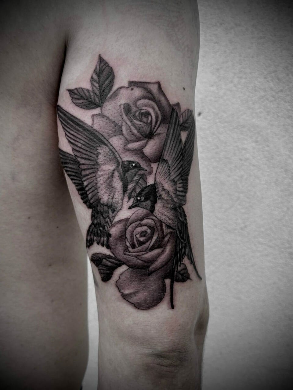 Black and grey birds with spread wings fly between roses on an upper arm.