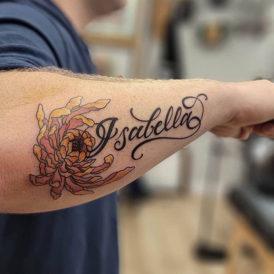 A neo traditional orange and pink chrysanthemum sits beside flowing black script on a forearm.