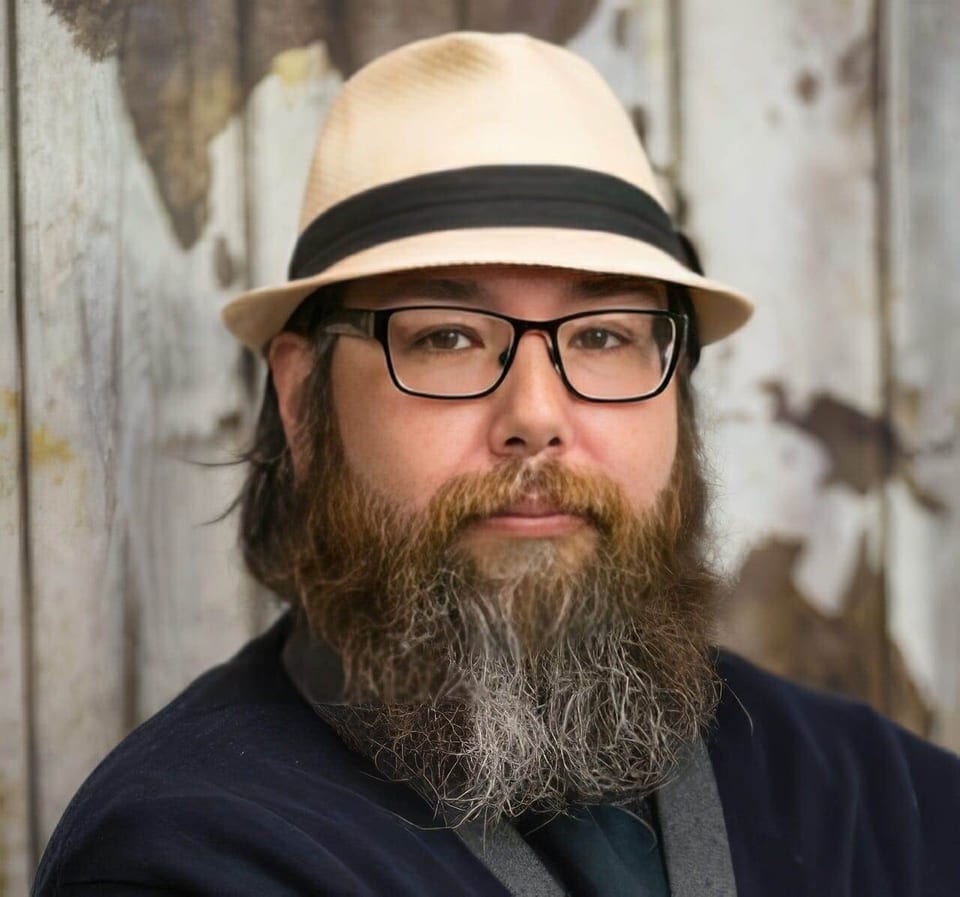Roger Solis wears a straw hat and glasses in front of a wooden wall.