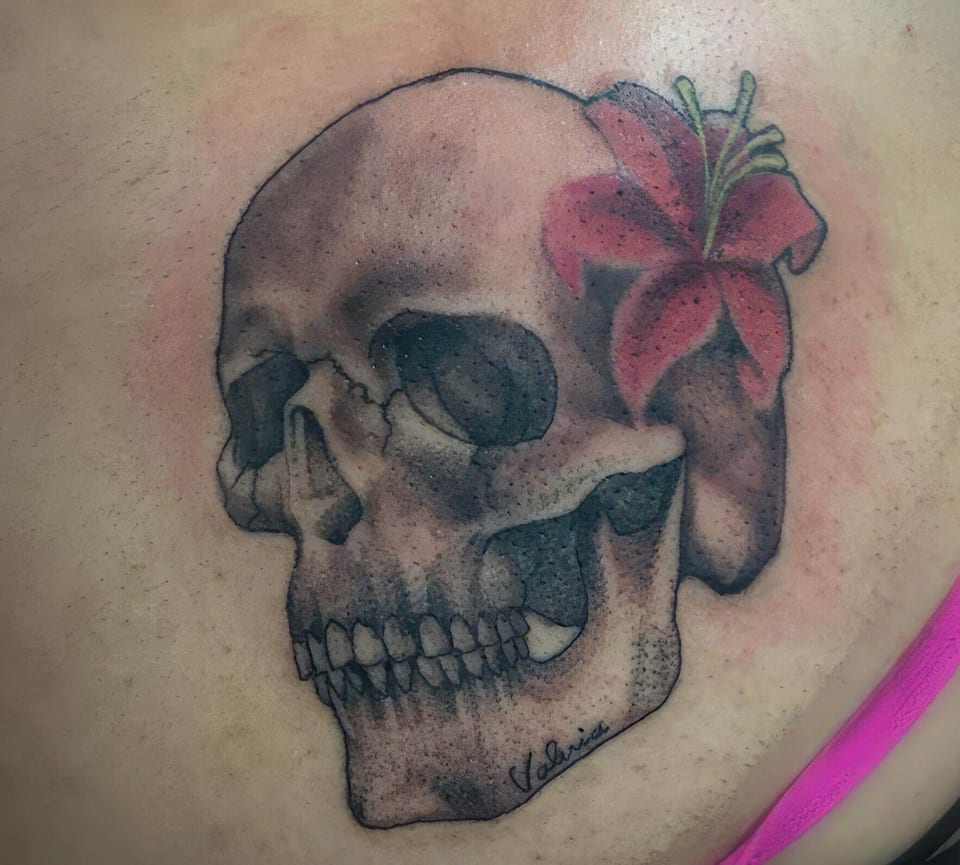 A black and grey skull with a pink lily sits on the shoulder blade.