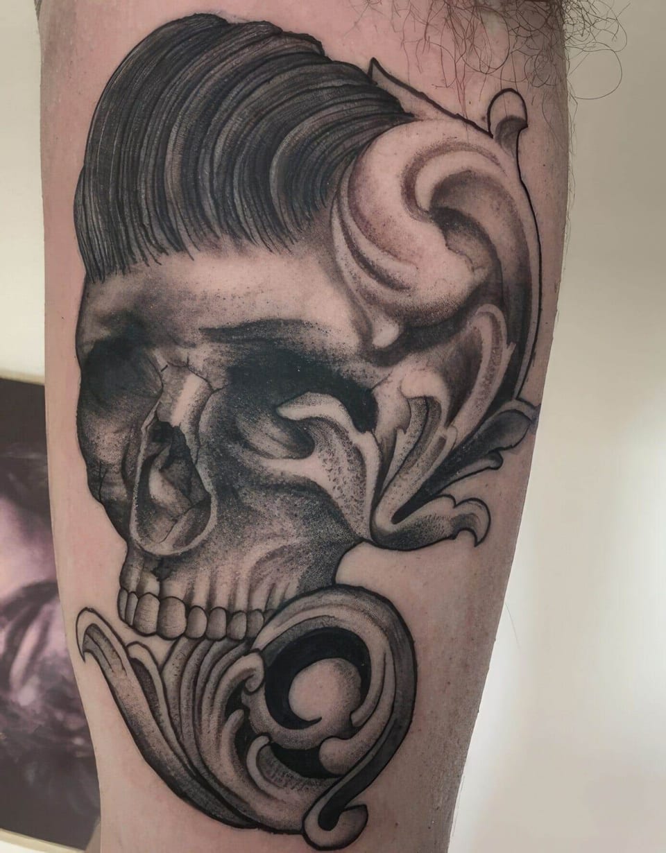A black and grey skull with a pompadour and scrolling ornament fills a thigh.