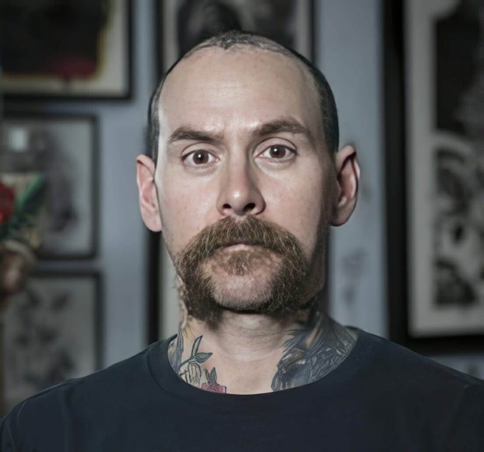 Skott Kautman faces the camera with neck tattoos visible in front of framed tattoo artwork.