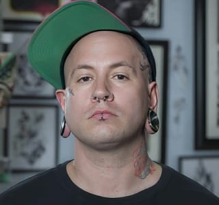 Arty faces the camera in a tilted green-brimmed cap before framed tattoo art.
