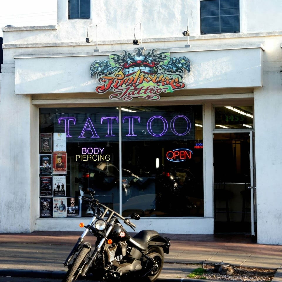 The Funhouse Tattoo storefront displays its illustrated sign and neon lettering behind a parked motorcycle.
