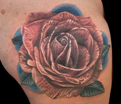 A realistic red rose with water droplets sits against blue shading on a thigh.
