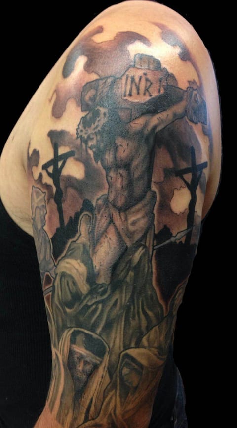 A black and grey realism crucifixion scene with kneeling figures covers the upper arm.