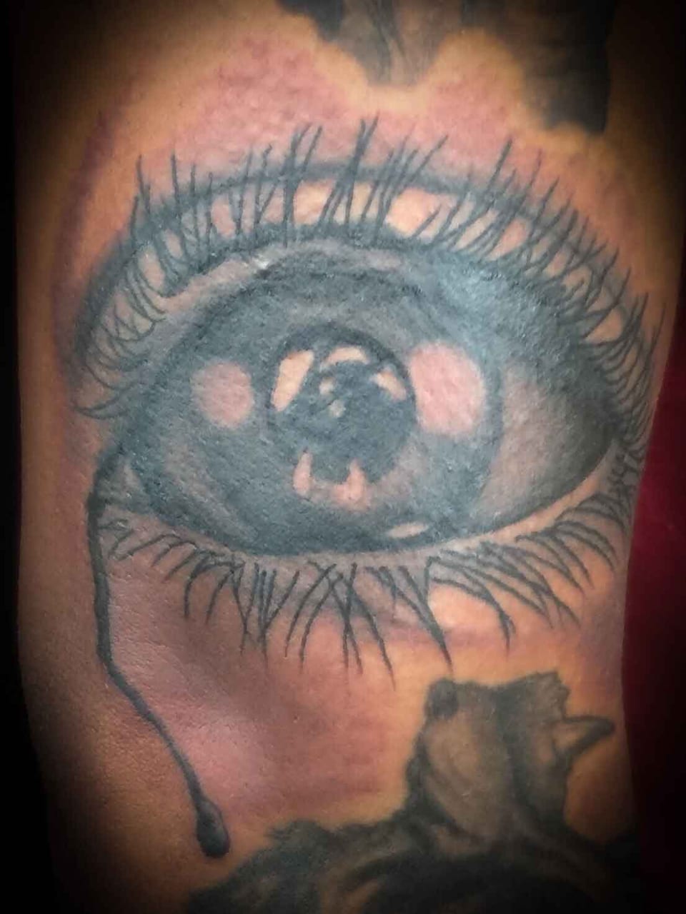 A black and grey realism eye with long lashes and a falling tear fills a close crop of tattooed skin.