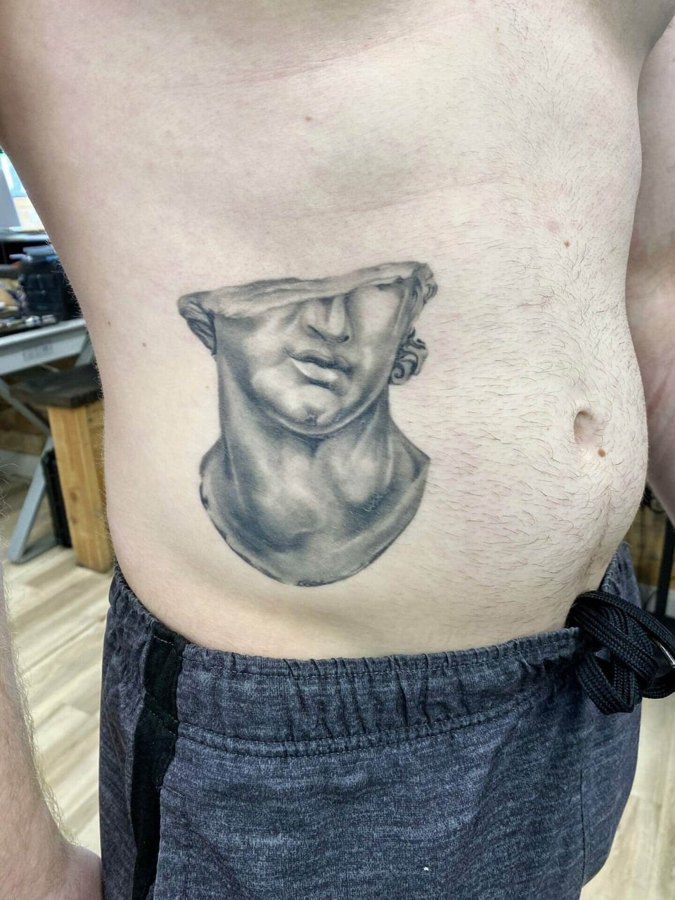 A black and grey realism fragment of a broken marble face sits on the ribs.