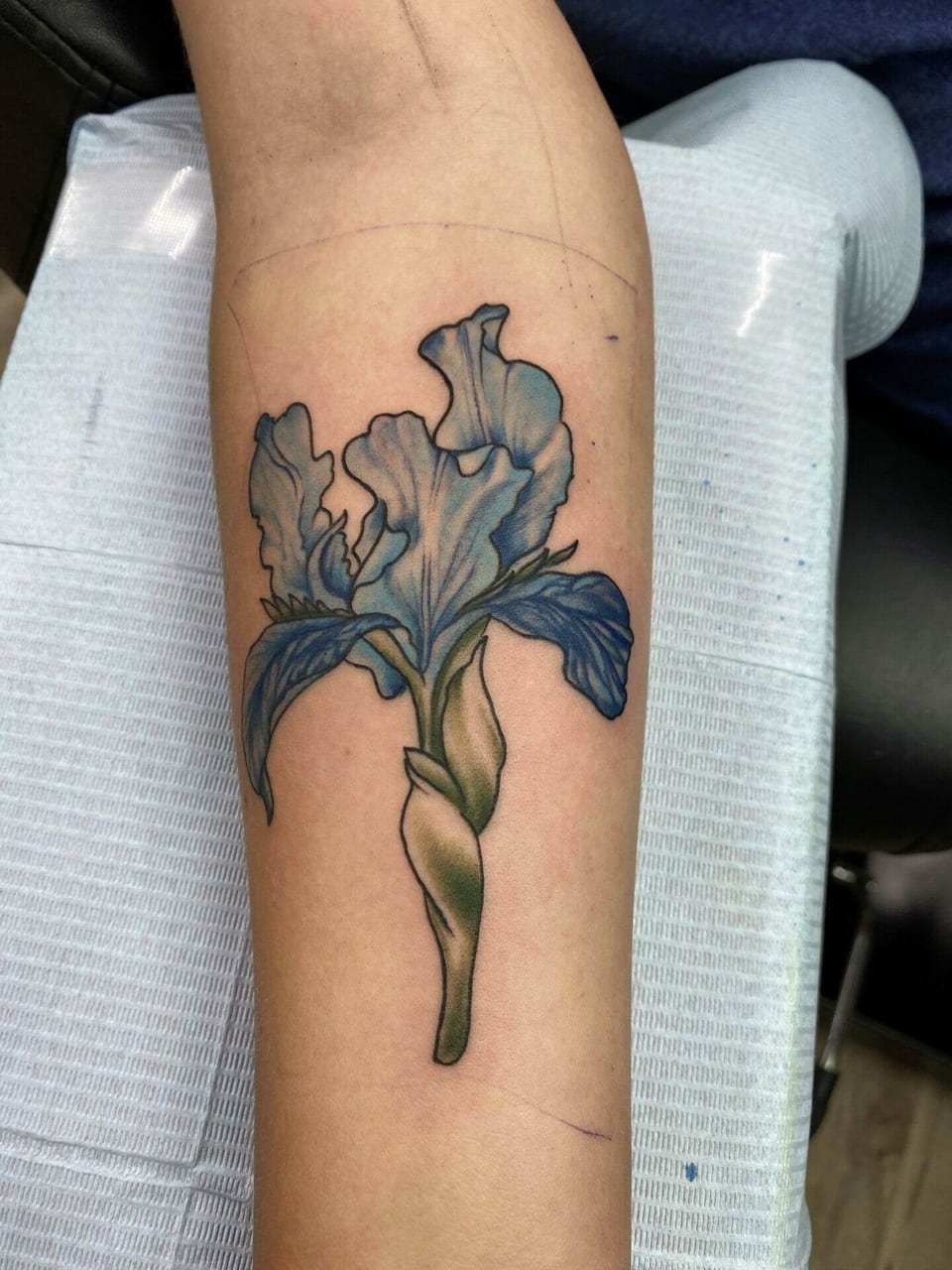 A realistic blue iris with a green stem and curling leaves covers the forearm.