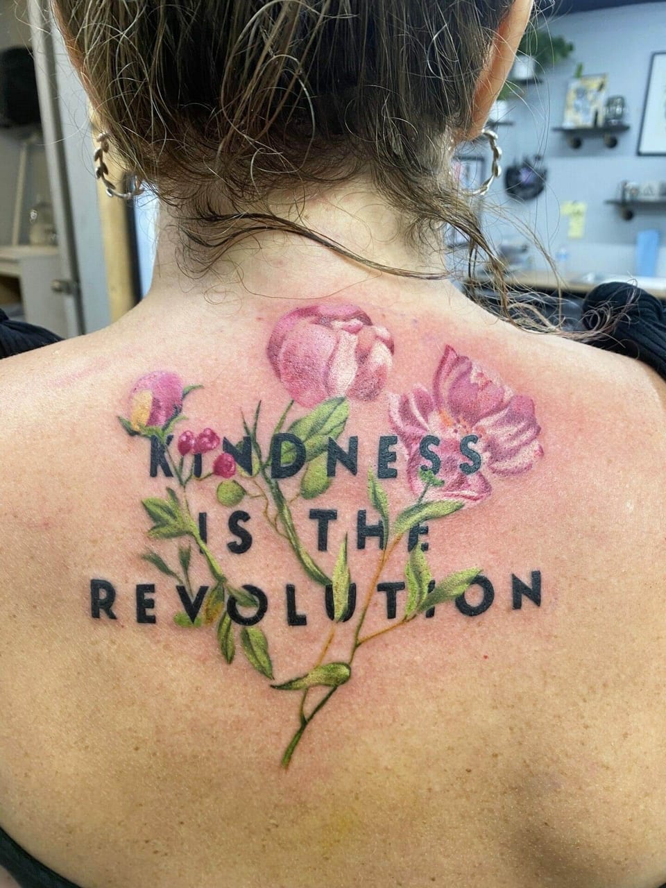Realistic pink flowers cross lettering reading "Kindness is the revolution" on the upper back.