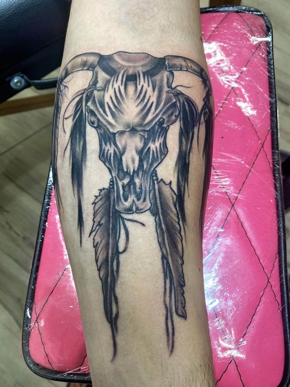 A black and grey illustrative horned animal skull with hanging feathers covers the forearm.