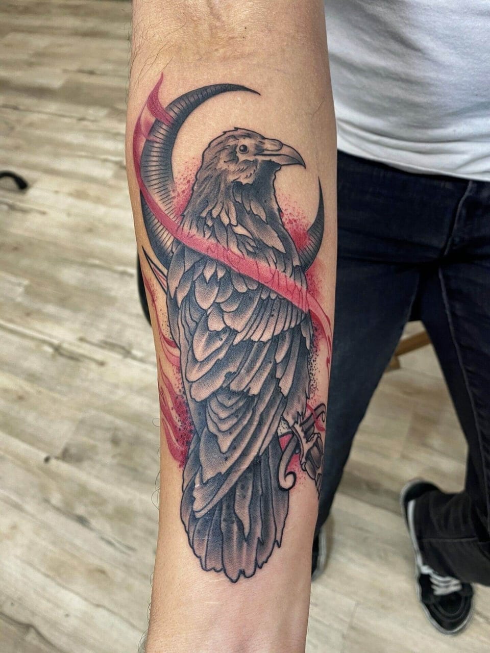 A neo-traditional black and grey raven sits within a crescent moon crossed by a red ribbon on the forearm.