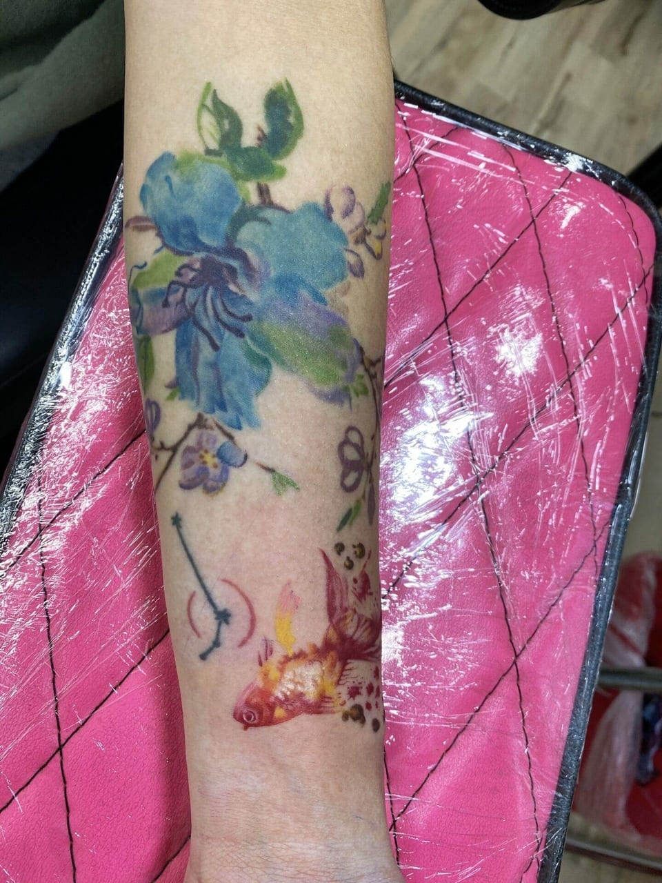Blue and green watercolor flowers appear above a red and yellow fish on a forearm.