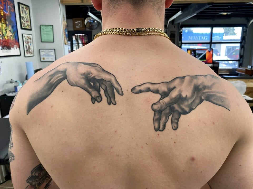 Black and grey realism hands reach toward one another across the upper back.